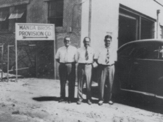 Black and white photo of three men next to a sign that says "Manda Bros Provision Co"