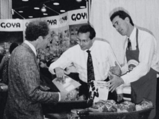 Black and white photo of two men showing an array of meats on a convention show floor